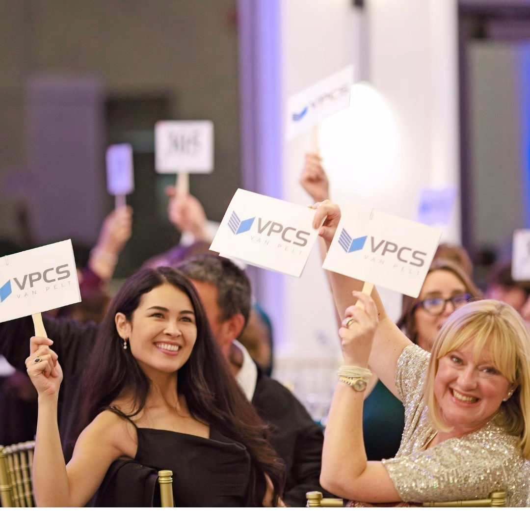 Two women smiling and holding up VPCS signs at a formal indoor event, surrounded by other people also raising similar signs, suggesting a lively or participatory atmosphere.