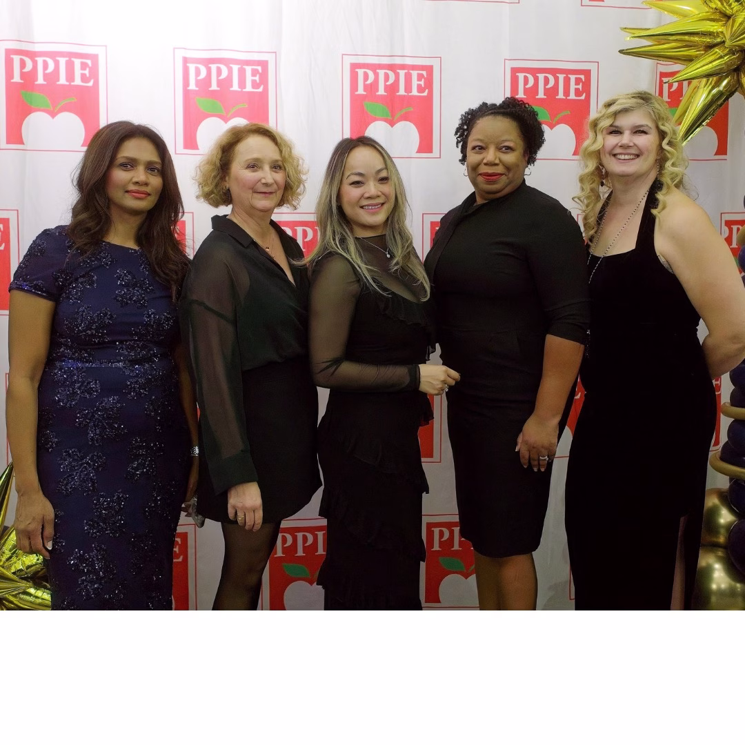 Five women dressed in semi-formal black attire stand together, smiling, in front of a backdrop with repeating red and white PPIE logos and decorative gold elements.