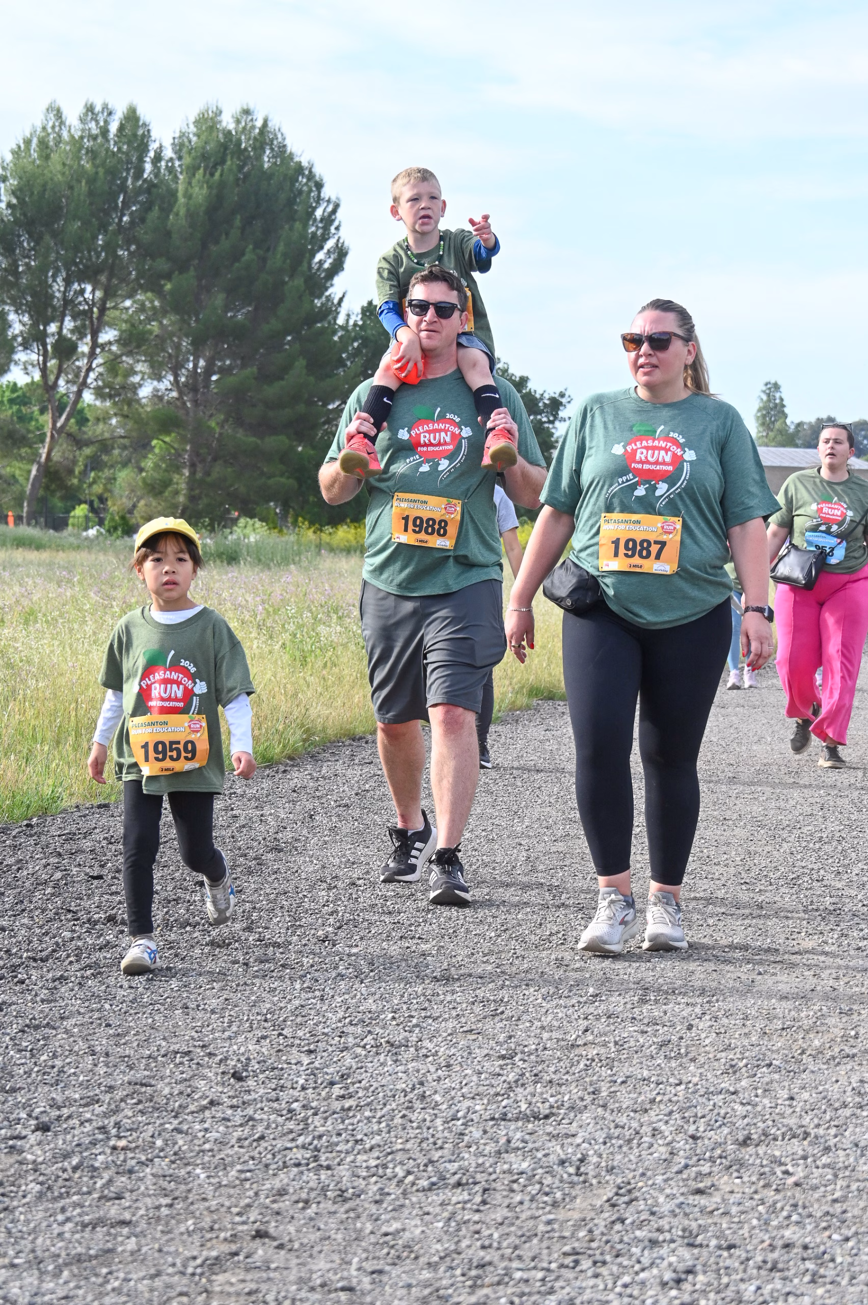 A family of four walks on a gravel path during a fun run event, all wearing green event shirts and numbered bibs. The man carries a child on his shoulders, while a woman and another child walk beside them. Trees and grass are in the background.