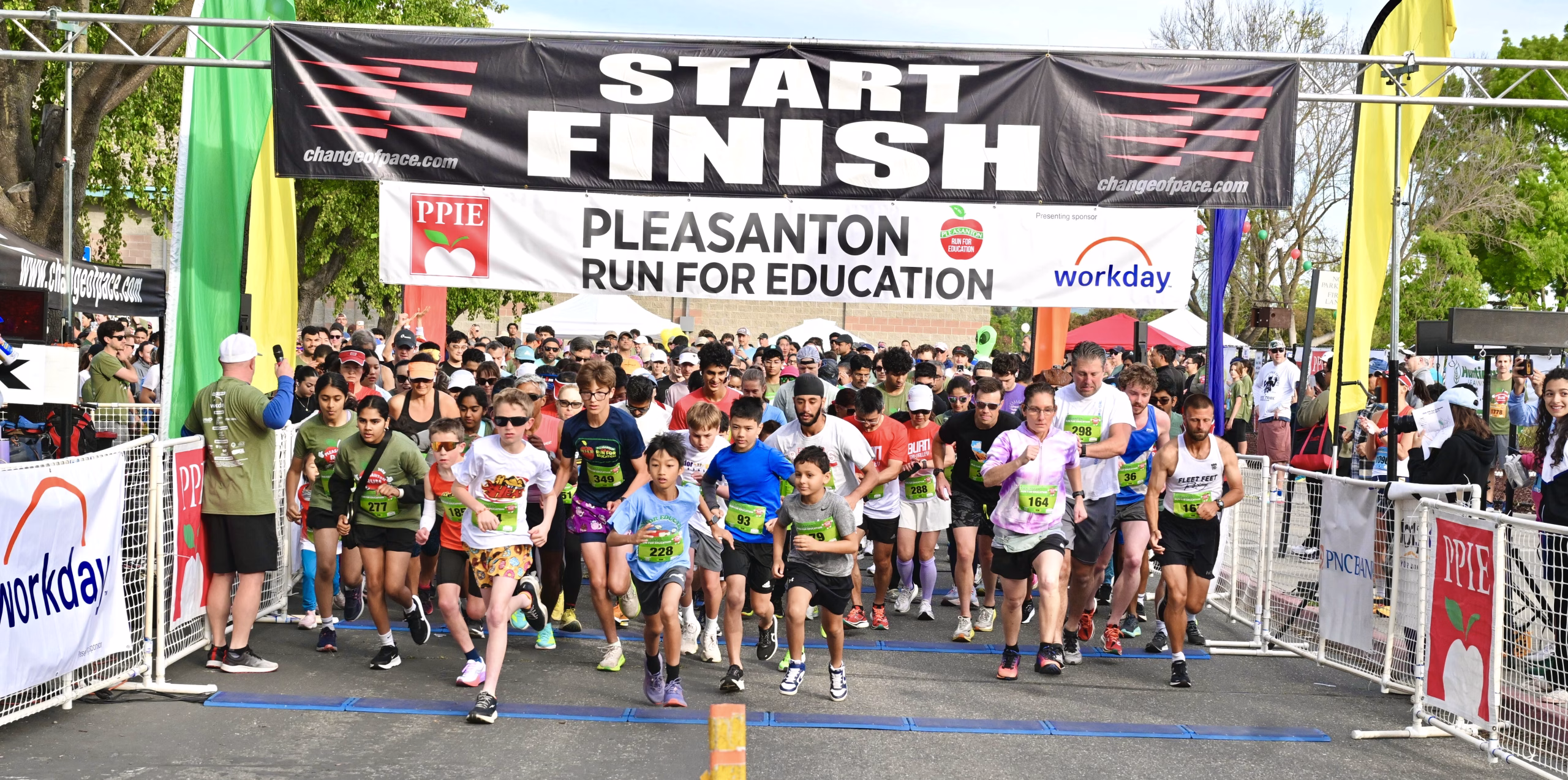 A large group of runners of various ages starts a race under a banner reading Start Finish, Pleasanton Run for Education. Spectators and volunteers watch from the sides on a sunny day.